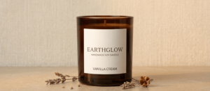 A handmade soy candle in an amber glass jar on a light wood surface. The minimalist beige label features the "EarthGlow" wordmark in serif typography and "Vanilla Cream" scent description.