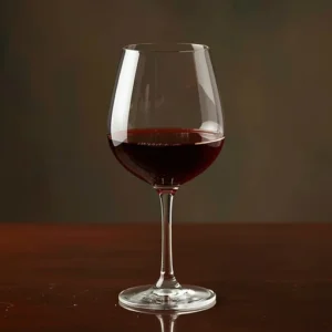Taça transparente cheia até a metade com vinho tinto, sobre mesa de madeira marrom.