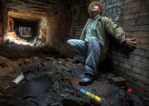 Photo of a man leaning against the wall of a tunnel strewn with bricks and trash. He looks upward, as if for escape.