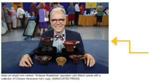 Photo of a commentator from Antiques Roadshow, sitting at a table, wearing a bow tie, suit, and dark glasses, smiling.